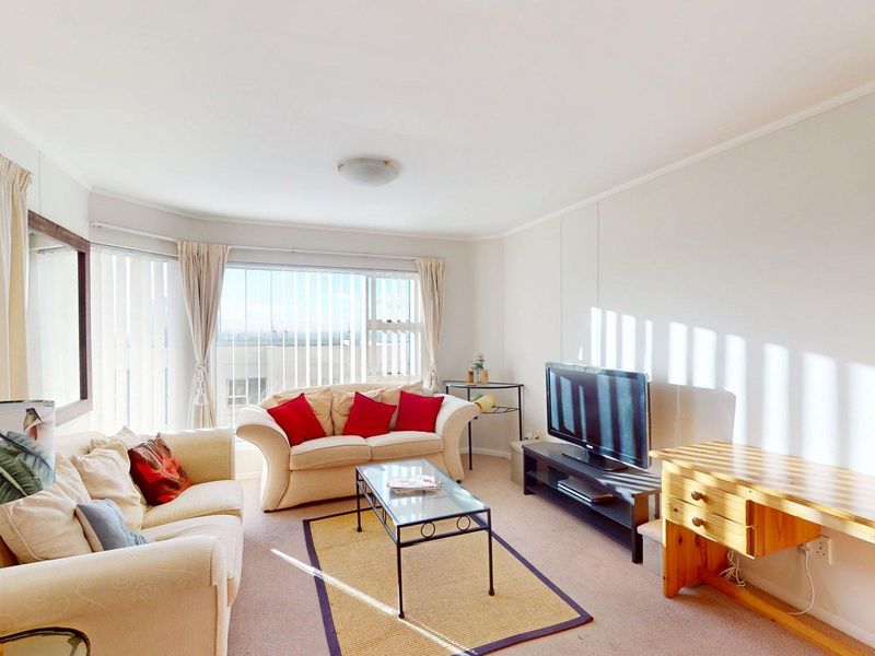 A bright living room featuring large windows with vertical blinds, neutral wall finishes, and carpeted flooring, offering a comfortable and well-lit space.