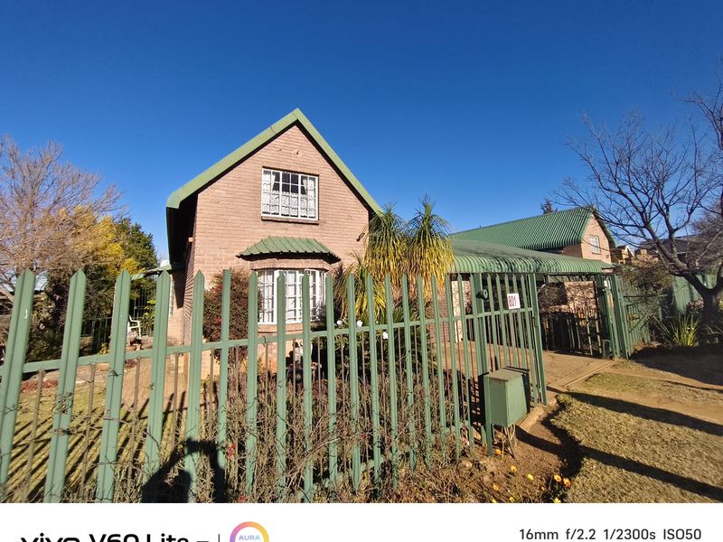 Front view of a brick house featuring a green metal roof, bay window, and secure metal perimeter fencing with an attached carport.