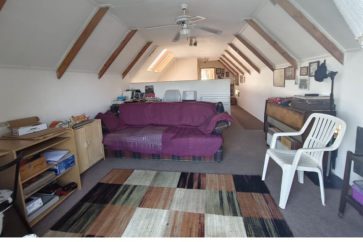 An attic-style living room featuring exposed timber rafters, a ceiling fan, and skylights, offering a unique architectural space with potential for modernization.
