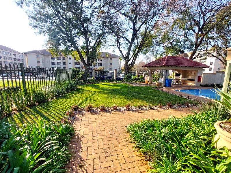 A well-maintained communal pool area featuring a brick-paved patio, a covered lapa, and landscaped garden surroundings within a secure apartment complex.