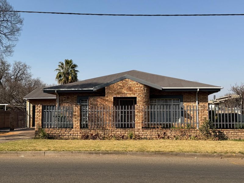 Front elevation of a single-story face brick house featuring a metal roof, secure perimeter fencing, and a paved driveway.