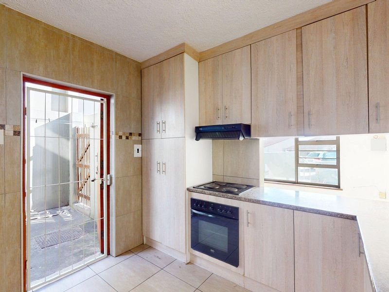 A functional kitchen featuring light wood-finish cabinetry, built-in oven and hob, tiled walls, and direct access to an outdoor area via a security gate.