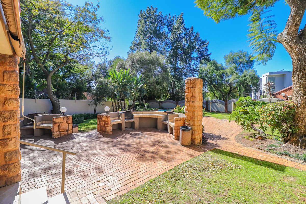 A well-maintained rear garden featuring a built-in brick braai area, stone masonry pillars, and extensive brick paving surrounded by mature trees and landscaping.