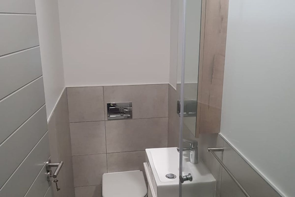A modern, compact bathroom featuring a wall-hung toilet, vanity basin with integrated storage, and a mirrored medicine cabinet with neutral tiled finishes.