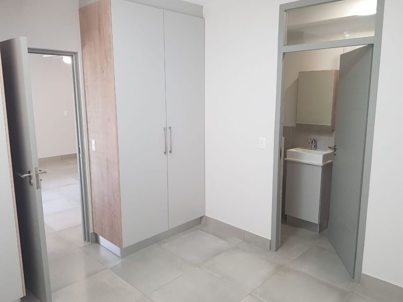 A clean, modern bedroom featuring built-in cupboards, neutral tiled flooring, and an adjoining en-suite bathroom with a vanity basin.
