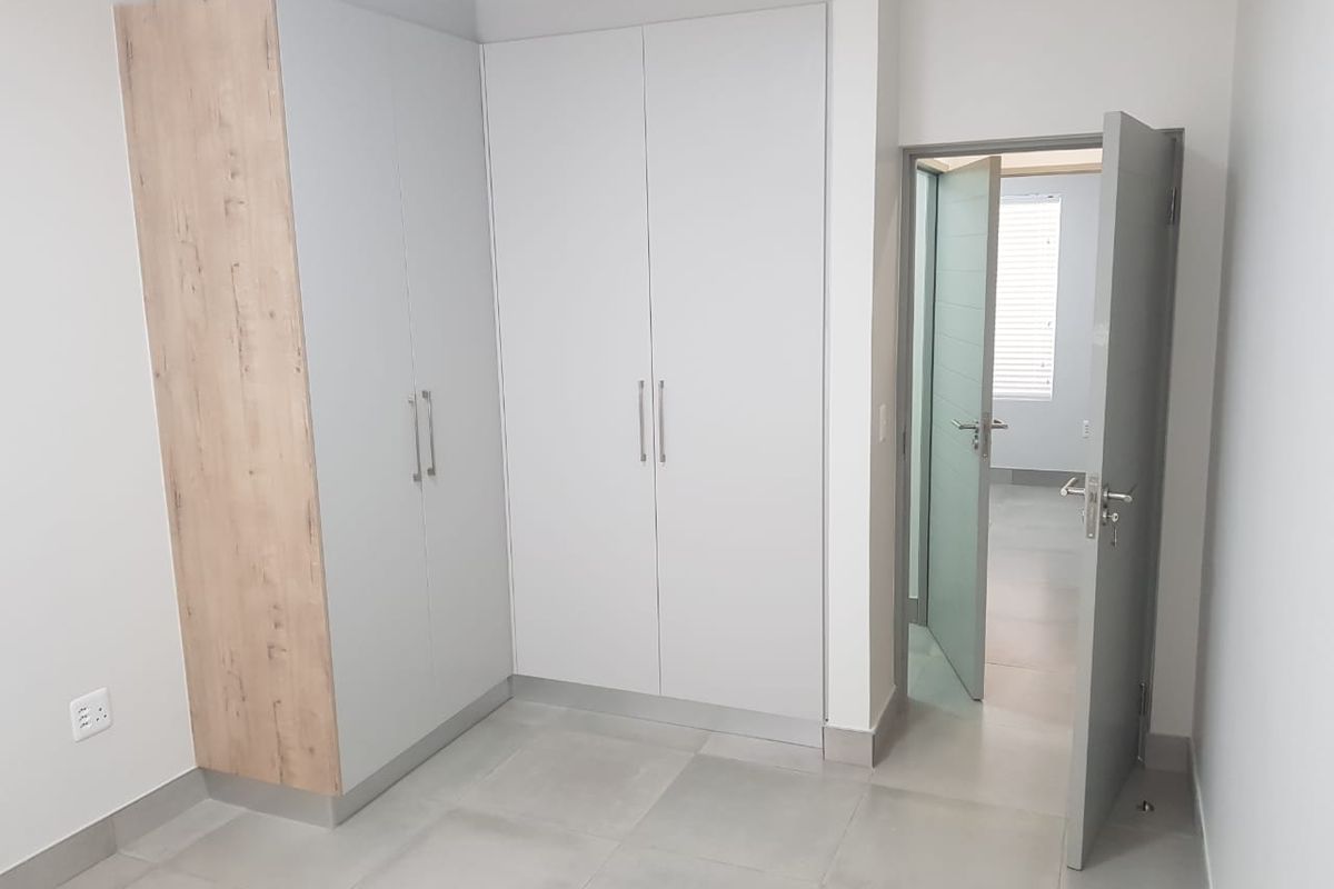 A clean, modern bedroom featuring neutral-toned built-in cupboards, large format floor tiles, and a contemporary internal door leading to an adjacent space.
