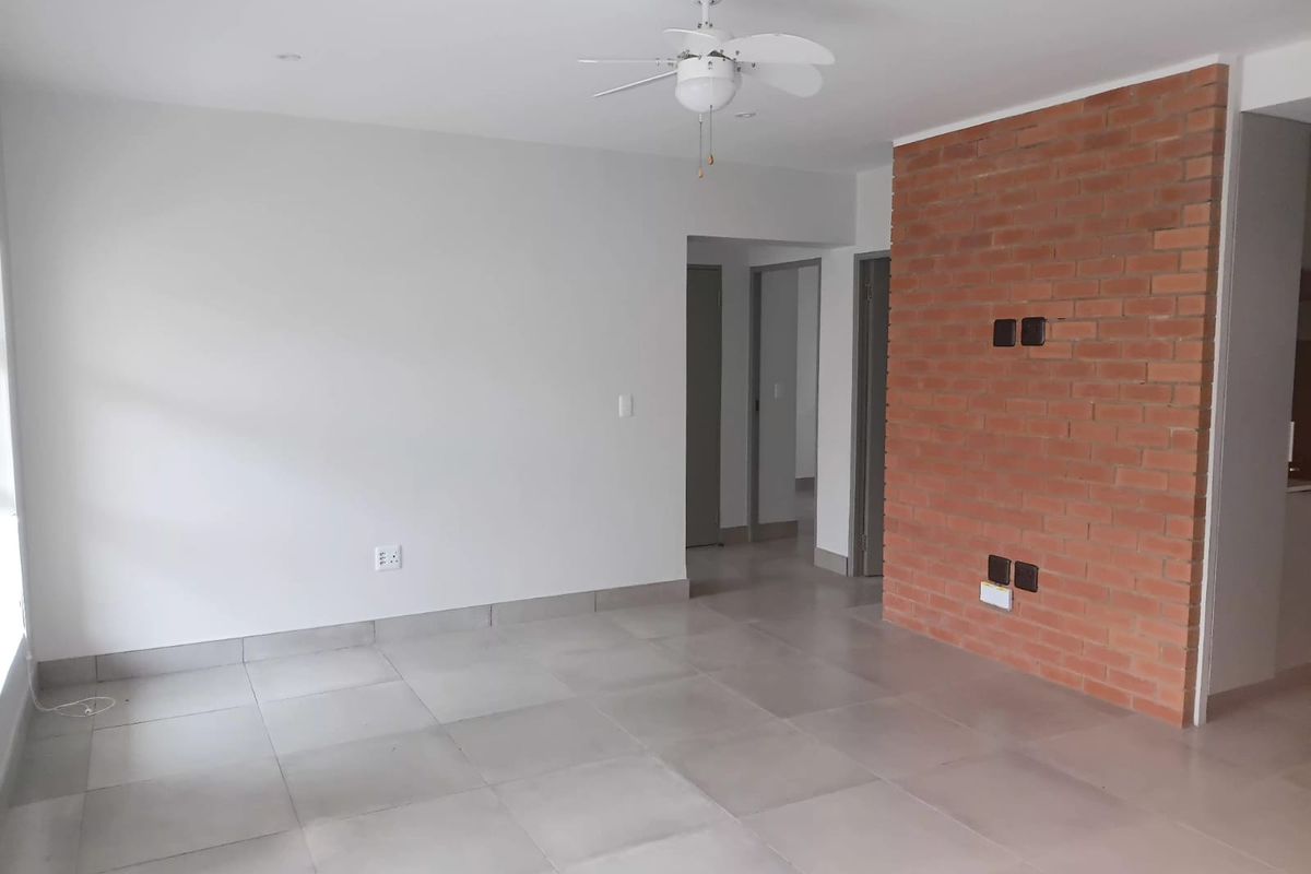 A bright, open-plan living area featuring neutral tiled flooring, a ceiling fan, and a prominent exposed brick feature wall.