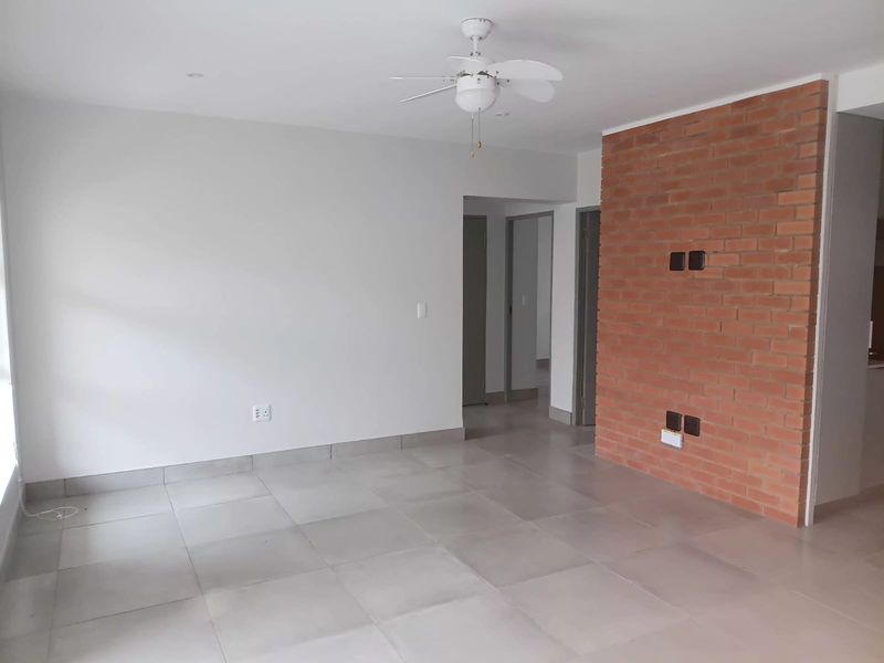 A bright, open-plan living area featuring neutral tiled flooring, a ceiling fan, and a prominent exposed brick feature wall.