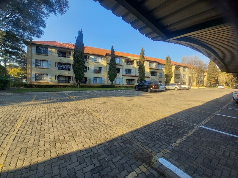 Exterior view of the apartment complex featuring a paved parking area, covered parking bays, and multi-story residential units with balconies.