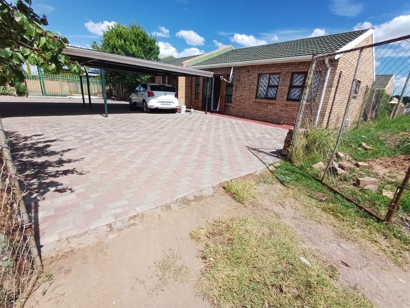 Front exterior view of a brick house featuring a paved driveway, covered carport, and perimeter fencing.