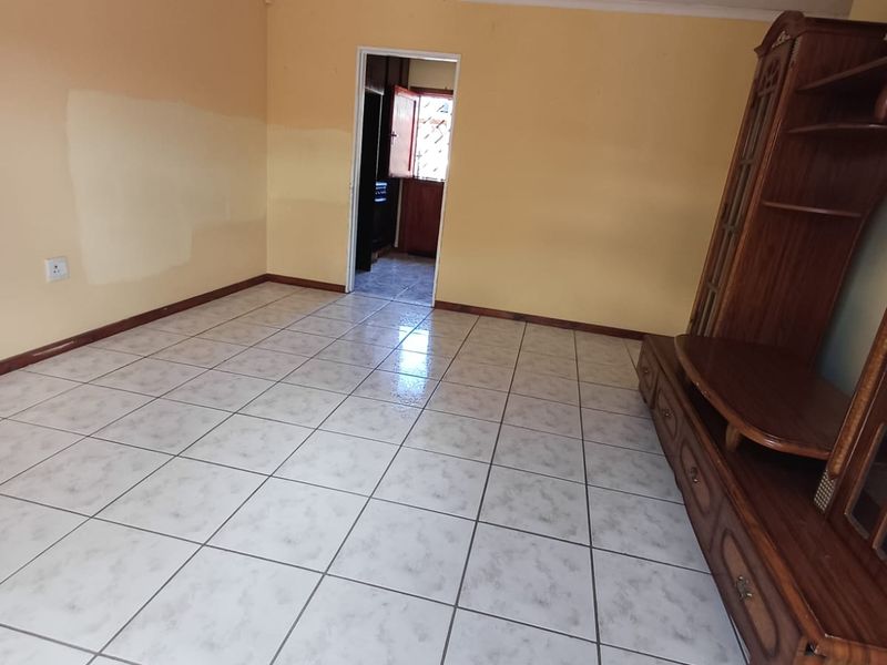 A living room featuring ceramic floor tiles and yellow painted walls with visible patch repairs, leading through to an adjacent kitchen area.