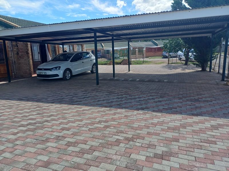A spacious outdoor carport area featuring paved flooring and a metal roof structure, providing covered parking space adjacent to the main house.