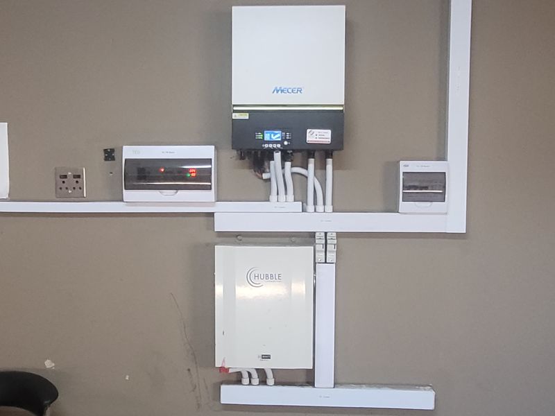A wall-mounted solar power system featuring a Mecer inverter and Hubble lithium battery with organized electrical distribution boards and conduit trunking.