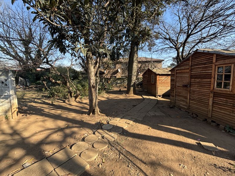 A spacious garden area featuring mature trees, a paved pathway, and a wooden storage shed structure.