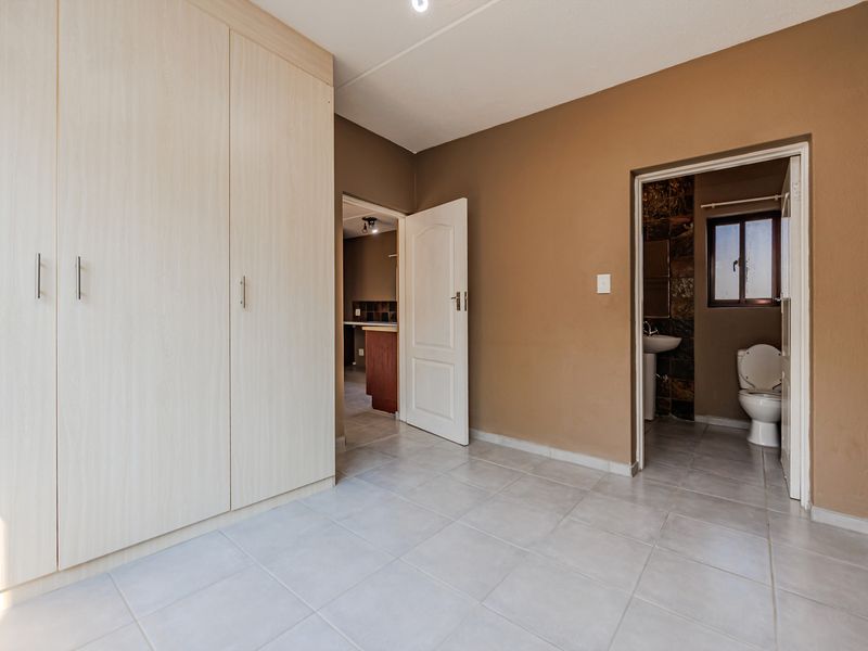 A spacious, empty bedroom featuring light-toned built-in cupboards, neutral tiled flooring, and direct access to an en-suite bathroom.