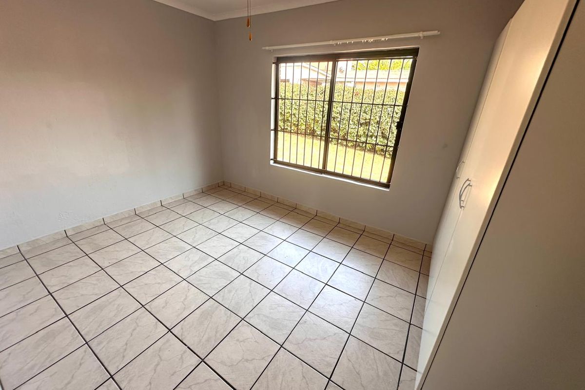 A clean, empty bedroom featuring neutral tiled flooring, a built-in wardrobe, and a window fitted with security bars overlooking the garden.