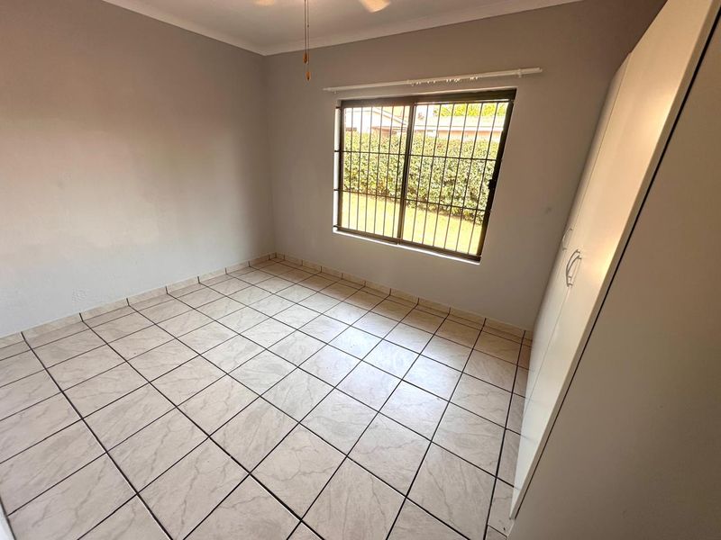 A clean, empty bedroom featuring neutral tiled flooring, a built-in wardrobe, and a window fitted with security bars overlooking the garden.