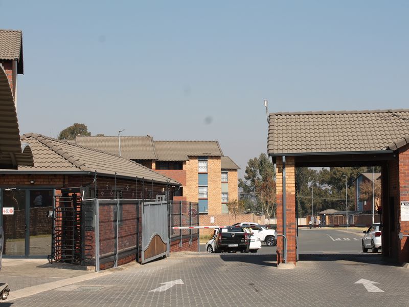 A view of the apartment complex entrance featuring a security gate, boom barrier, and paved driveway access.