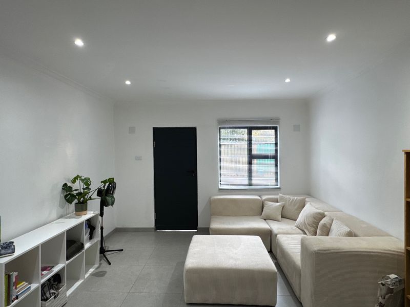 A bright, modern living room featuring neutral tiled flooring, recessed downlighting, and a large window with fitted venetian blinds.