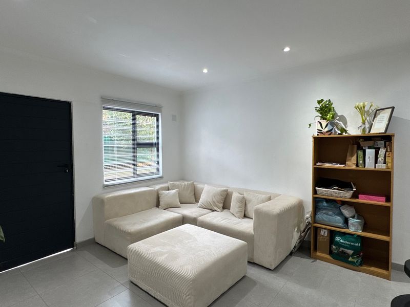 A bright and modern living room featuring neutral tiled flooring, recessed downlighting, and a large window with fitted venetian blinds.