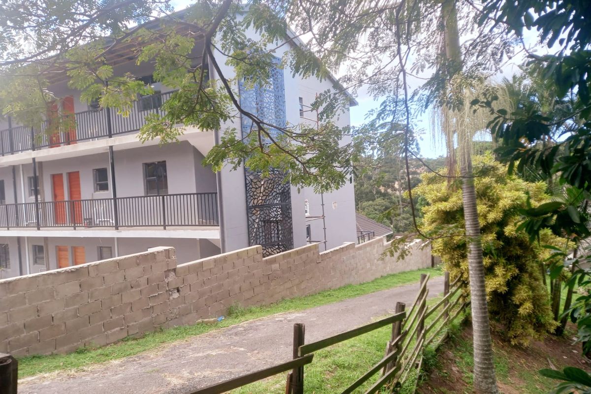 Exterior view of an apartment building featuring metal balcony railings, a decorative screen, and a concrete block boundary wall along the driveway.