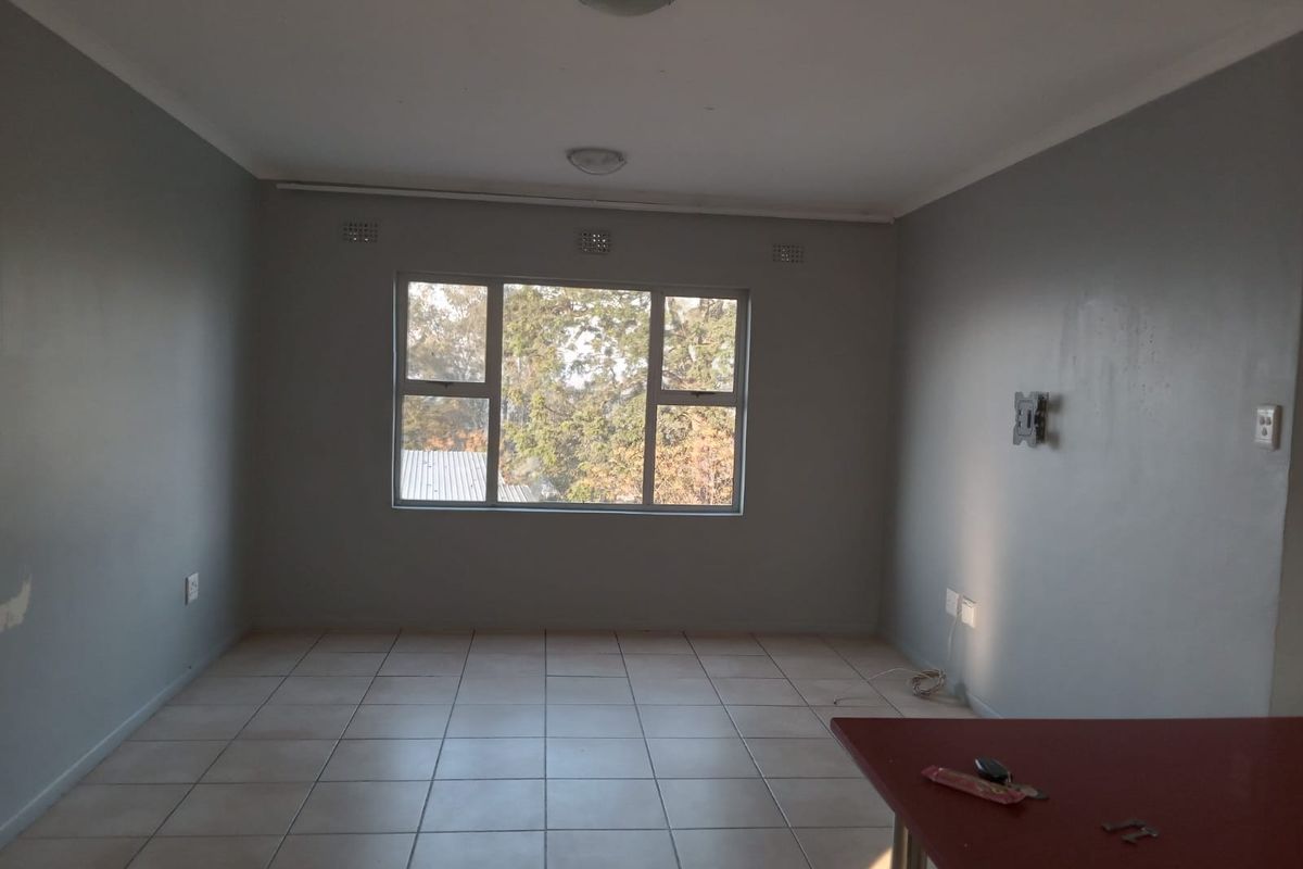 A spacious, empty living area featuring neutral grey walls, tiled flooring, and large windows providing natural light.