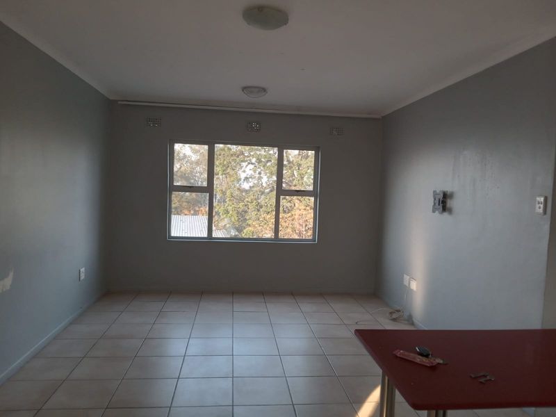 A spacious, empty living area featuring neutral grey walls, tiled flooring, and large windows providing natural light.