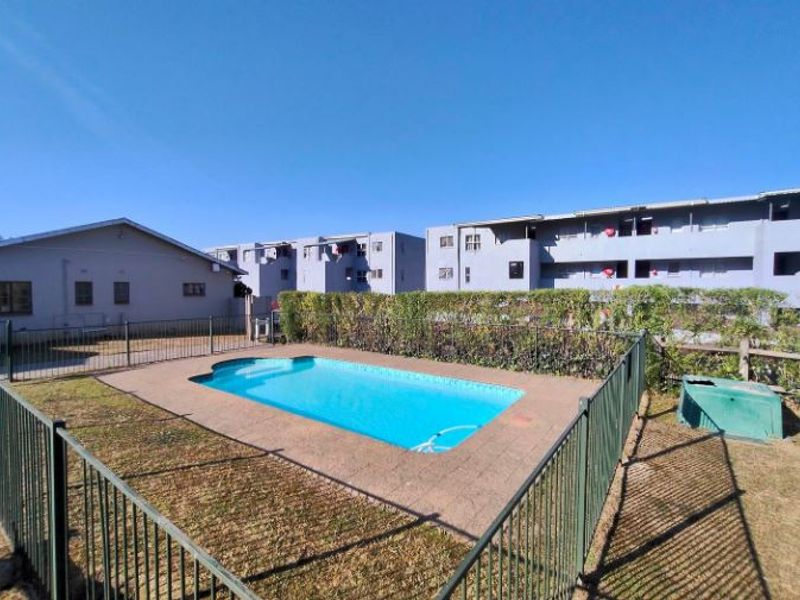 Communal swimming pool area featuring a paved surround and metal perimeter fencing, set within a residential apartment complex.