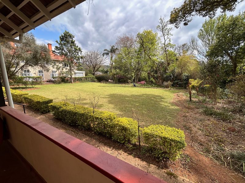A spacious, well-maintained garden featuring a large lawn, established hedging, and mature trees, viewed from a covered patio area.