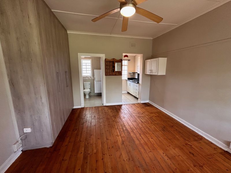 Spacious bedroom featuring polished timber flooring, built-in cupboards, and a ceiling fan, with direct access to an adjoining bathroom and kitchen area.