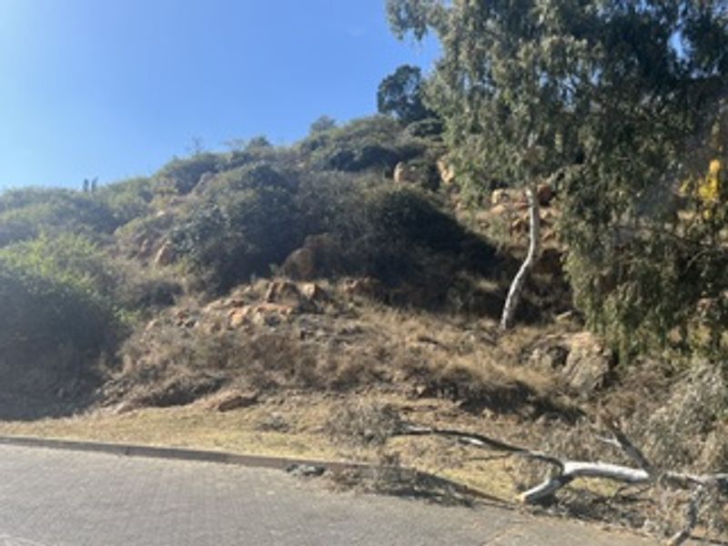 A view of a sloped, undeveloped residential plot featuring natural rocky terrain and established trees, accessible from the paved street.