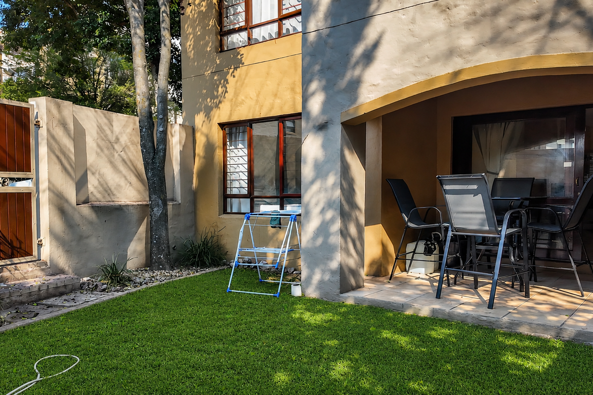 A rear exterior view of an apartment featuring a covered patio with tiled flooring, sliding glass doors, and a small private garden area.