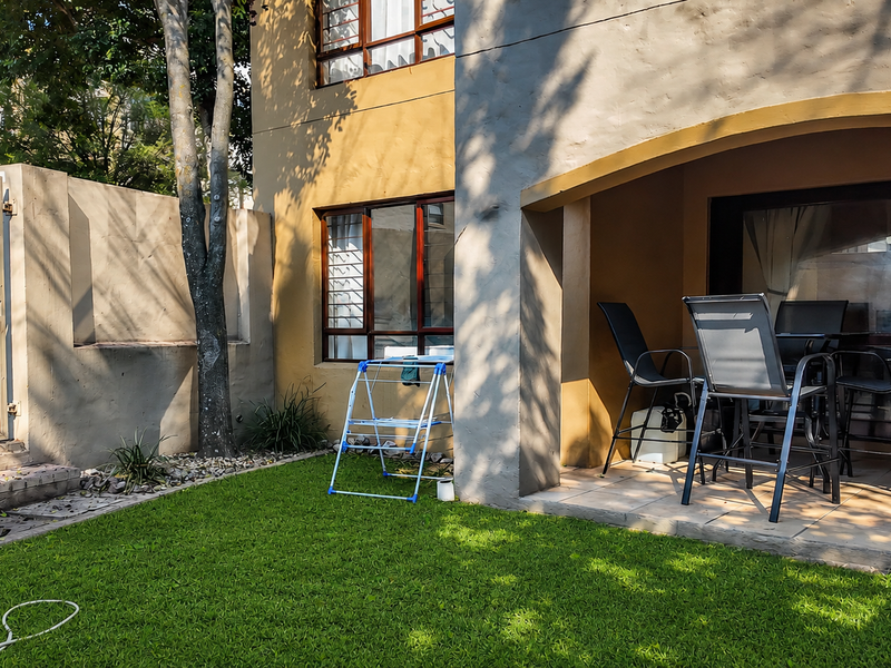 A rear exterior view of an apartment featuring a covered patio with tiled flooring, sliding glass doors, and a small private garden area.