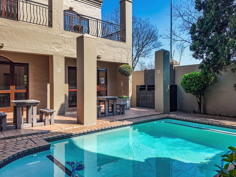 Outdoor pool area featuring a tiled patio, built-in braai, and access to the apartment building with a balcony above.
