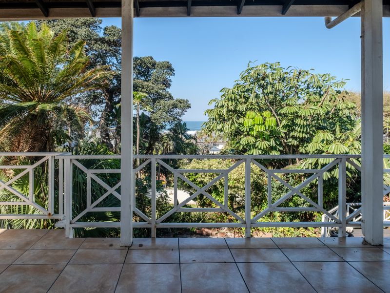 A covered balcony featuring tiled flooring and a timber balustrade, offering a scenic view of the surrounding garden and distant ocean.