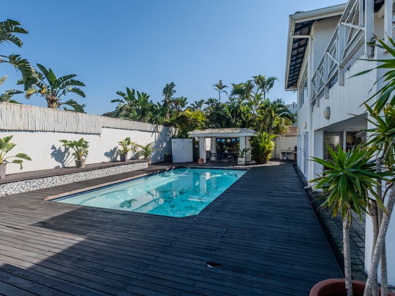 A spacious outdoor pool area featuring a swimming pool surrounded by timber decking, a covered patio, and a boundary wall with lush garden landscaping.