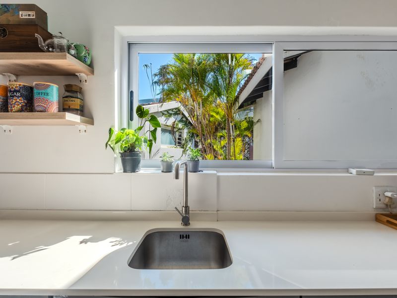 A bright kitchen area featuring a clean stone countertop, stainless steel undermount sink, modern mixer tap, and floating timber shelving with a garden view.