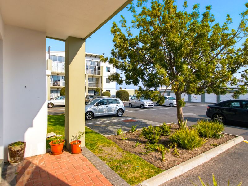 A covered patio area with brick paving and a concrete support pillar, overlooking the apartment complex parking area and landscaped garden.
