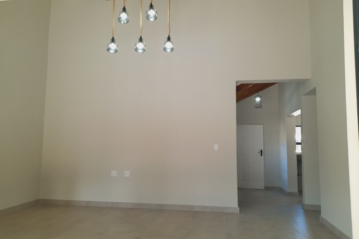 A bright, empty living area featuring neutral tiled flooring, high ceilings with modern pendant lighting, and access to an adjoining hallway.