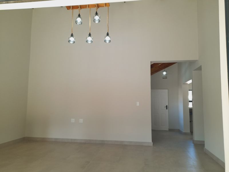 A bright, empty living area featuring neutral tiled flooring, high ceilings with modern pendant lighting, and access to an adjoining hallway.