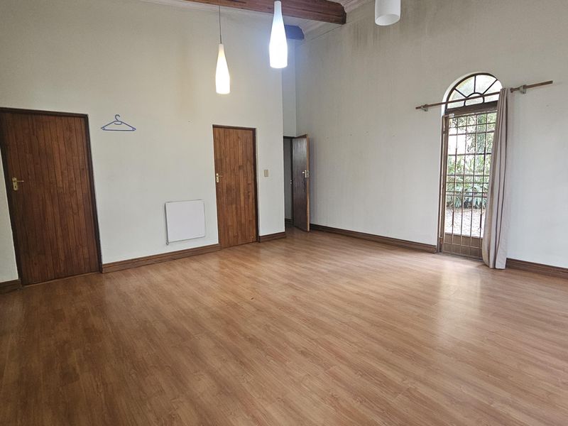 An empty living room featuring laminate flooring, timber doors, and an arched glass door leading to the exterior, with pendant lighting and exposed ceiling beams.