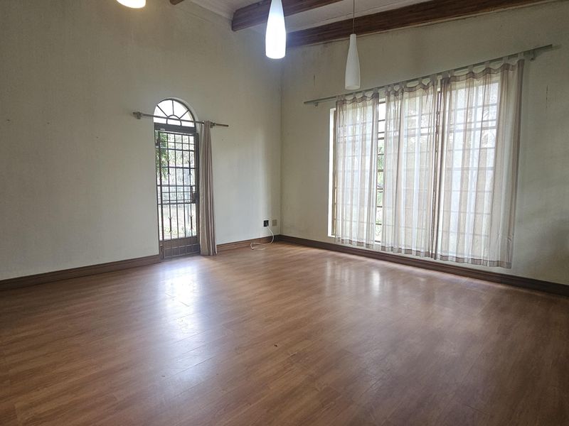 Spacious living room featuring laminate flooring, exposed timber ceiling beams, and natural light through a large window and arched security door.