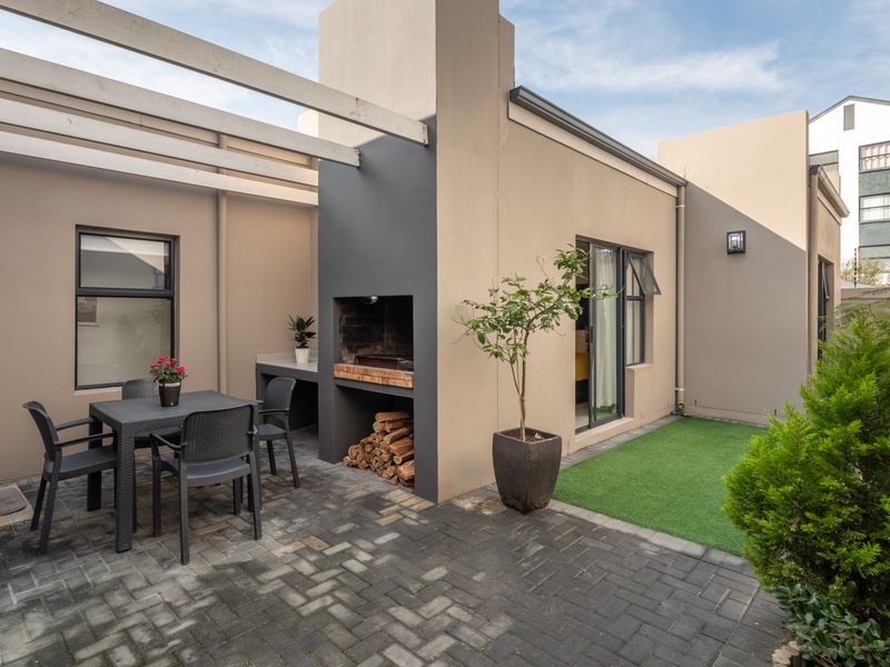 A well-maintained outdoor patio area featuring a built-in braai, pergola structure, paved flooring, and a small artificial lawn section.