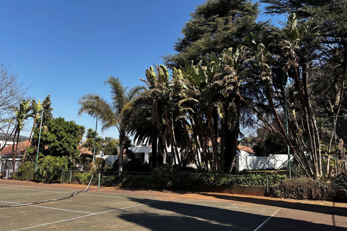 A private tennis court surrounded by mature trees and landscaping, featuring a worn surface and perimeter fencing.