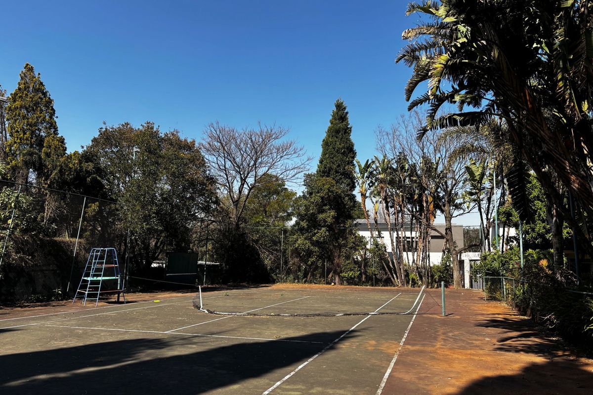 A private outdoor tennis court featuring a chain-link fence enclosure and a worn playing surface surrounded by mature trees.
