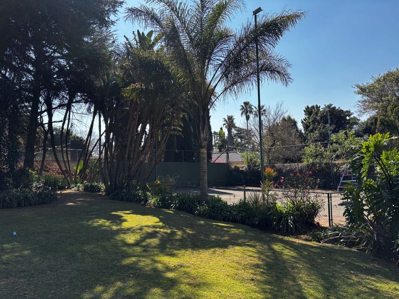 A private outdoor tennis court surrounded by mature trees and landscaped garden areas, featuring perimeter fencing and a floodlight.