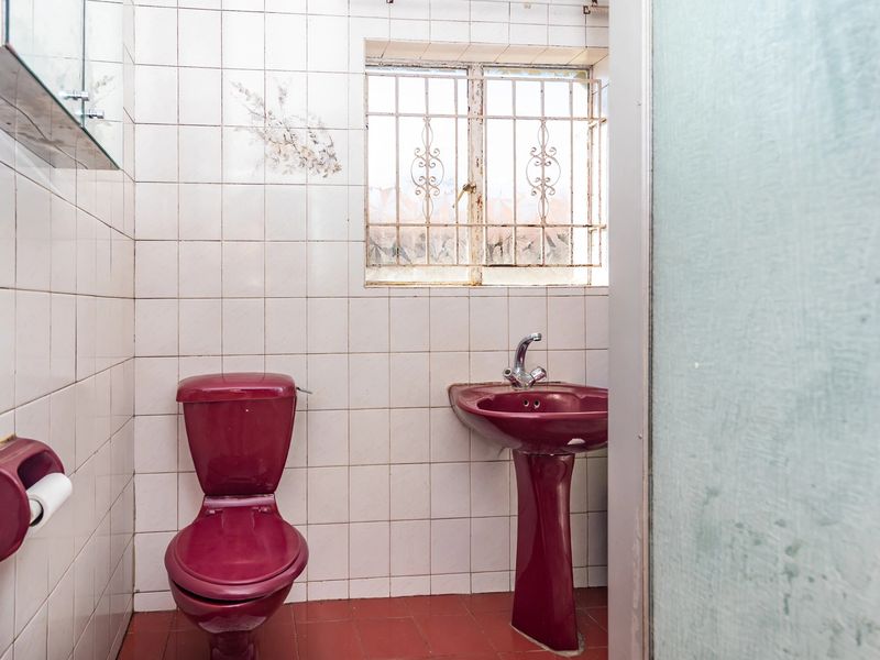 A dated bathroom featuring burgundy sanitaryware, white wall tiles, and a window with security bars.