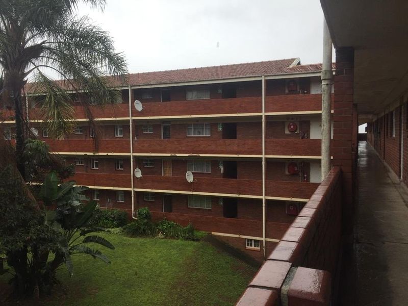 Exterior view of a multi-story brick apartment building featuring external walkways, tiled roofing, and communal garden space.
