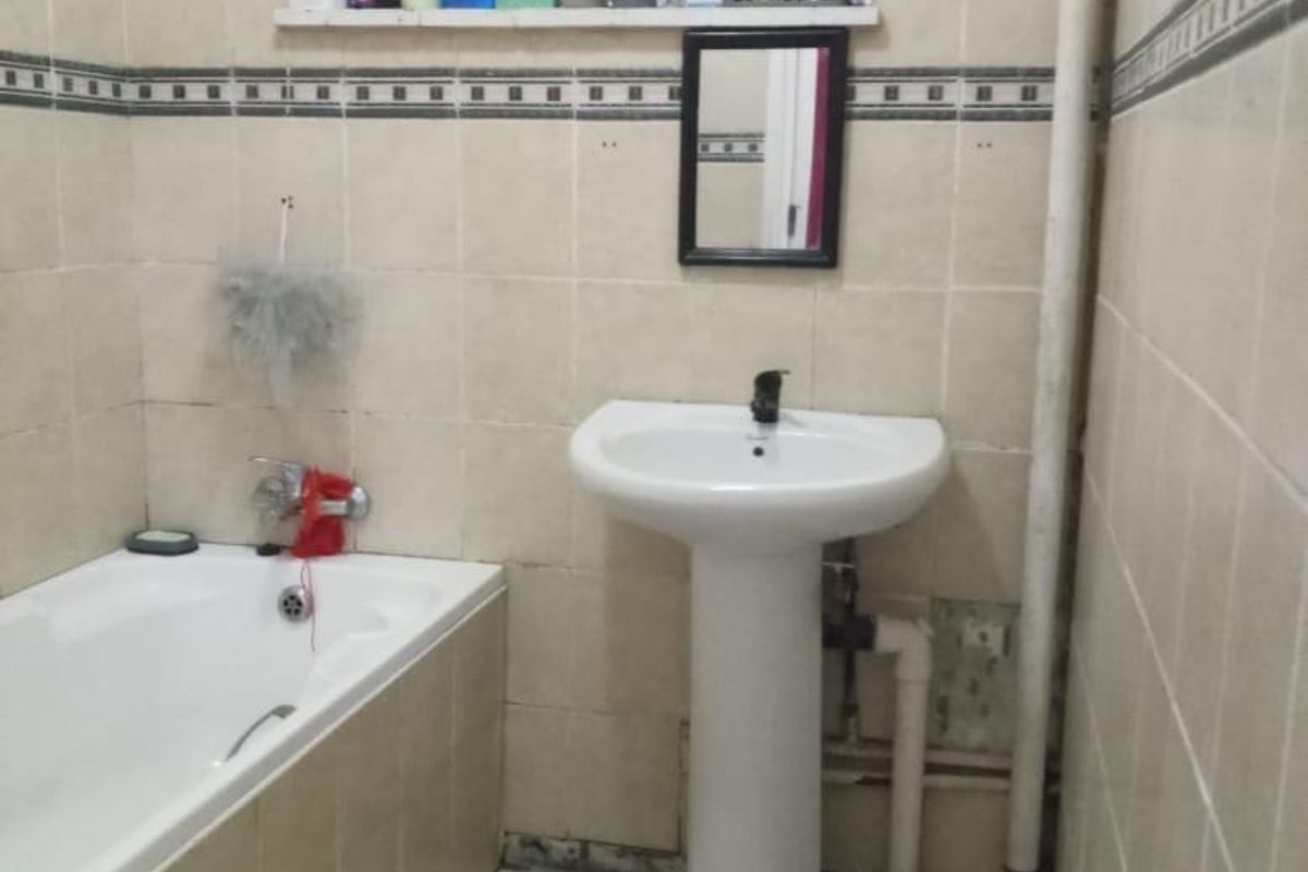 A dated bathroom featuring a pedestal basin, built-in bathtub, and tiled finishes, currently showing signs of wear and exposed plumbing.