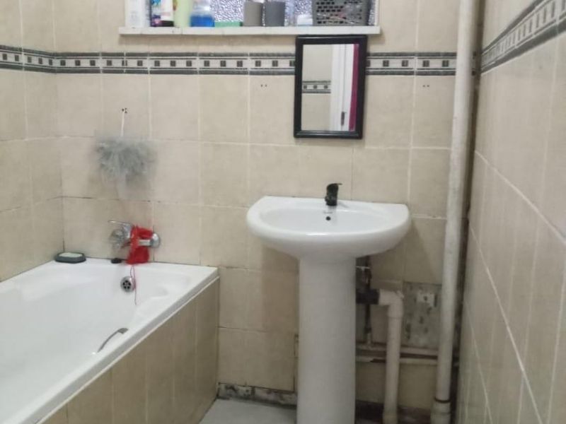 A dated bathroom featuring a pedestal basin, built-in bathtub, and tiled finishes, currently showing signs of wear and exposed plumbing.